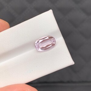 Puede incluir: Una gema ovalada de color púrpura claro, exhibida sobre una superficie blanca. La gema tiene una superficie facetada, que refleja la luz. El fondo es un material oscuro y texturizado. La gema está sujeta por los dedos de una persona.