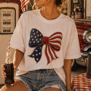 Könnte beinhalten: Weißes T-Shirt mit einem patriotischen Schleifendesign in Rot, Weiß und Blau, mit Sternen auf den blauen Abschnitten. Das Shirt wird mit Jeansshorts getragen. Eine alte Zapfsäule und ein Oldtimer sind im Hintergrund.