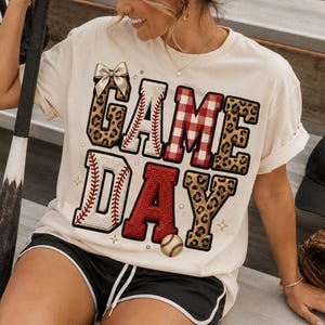 Puede incluir: Camiseta color crema con las palabras "GAME DAY" en un diseño temático de béisbol. Las letras presentan costuras de béisbol, estampado de leopardo y patrones de cuadros rojos. También se ve una gorra y un guante de béisbol.
