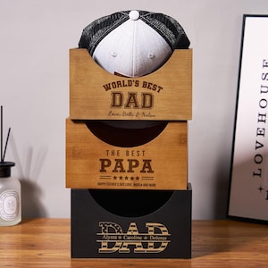 May include: Three wooden boxes with engraved messages for Dad. The top box is brown and has the text "World's Best Dad Love, Bella & Nolan". The middle box is brown and has the text "The Best Papa ***** Happy Father's Day Love, Melia and Mom". The bottom box is black and has the text "Dad Alyssa & Caroline & Dolores". A black and white baseball cap sits on top of the boxes.