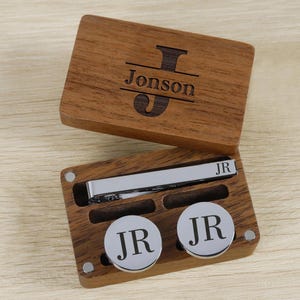 May include: A wooden box with a hinged lid, engraved with the name "Jonson" and a stylized "J". Inside, a silver tie clip and two circular cufflinks, each with "JR" engraved. The box and accessories are displayed on a light wood surface.