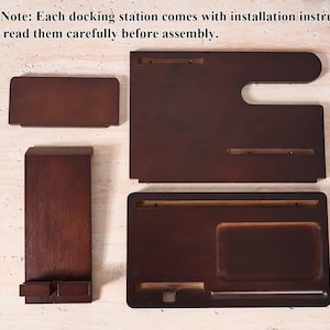 May include: A disassembled wooden docking station with multiple pieces. The pieces are various shapes and sizes, including a rectangular phone holder, a tray, and a larger piece with a curved edge. The wood has a dark brown finish. The text "Please Note: Each docking station comes with installation instructions, please read them carefully before assembly." is visible.
