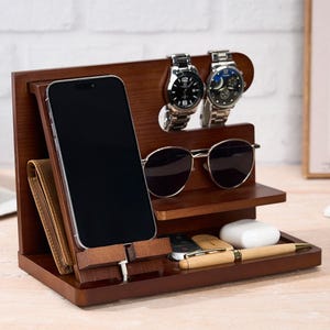 May include: A dark brown wooden desk organizer with compartments for a phone, wallet, sunglasses, watches, pen, keys, and earbuds. The organizer has a modern design and is made of wood.