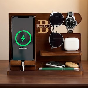 May include: A dark wood charging station with a phone displaying a charging symbol. The station holds sunglasses, a watch, and other accessories. The name "Bradford" is displayed in gold lettering.