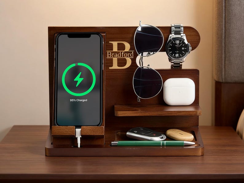 May include: A dark wood charging station with a phone displaying a charging symbol. The station holds sunglasses, a watch, and other accessories. The name "Bradford" is displayed in gold lettering.