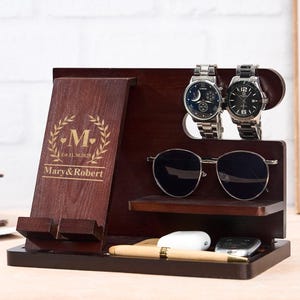 May include: A dark brown wooden organizer with a phone holder, watch holders, and a shelf for sunglasses. The organizer has the names "Mary & Robert" and the letter "M" with a laurel wreath design.