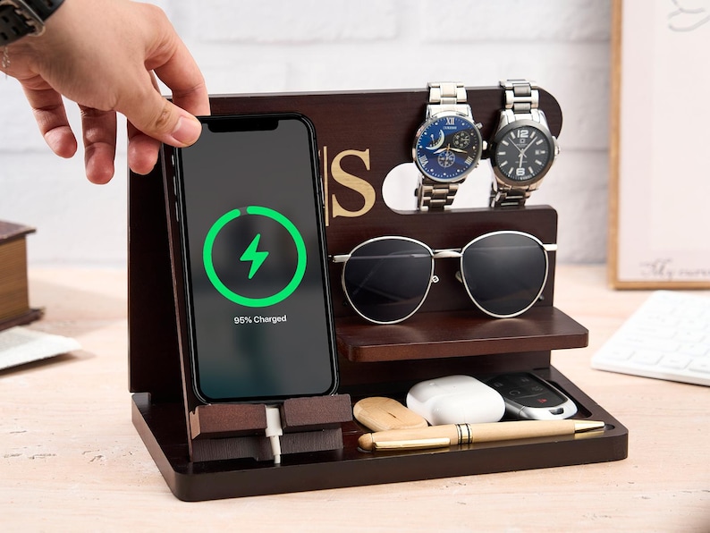 May include: A dark brown wooden charging station with a phone displaying a charging symbol. The station holds two watches, sunglasses, a pen, and other small items. The phone shows 95% charged.