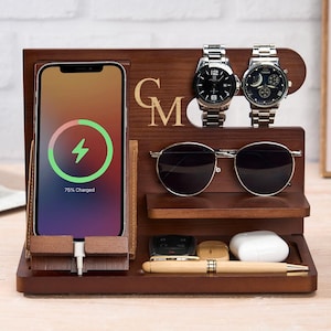 Könnte beinhalten: Eine hölzerne Ladestation mit einem Telefon, das ein Ladesymbol anzeigt. Sie verfügt über einen Uhrenhalter mit zwei silbernen Uhren, ein Regal für Sonnenbrillen und eine Ablage für kleine Gegenstände. Die Initialen "GM" sind eingraviert.