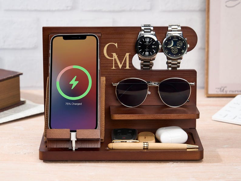 May include: A wooden charging station with a phone displaying a charging symbol. It features a watch holder with two silver watches, a sunglasses shelf, and a tray for small items. The initials "GM" are engraved on the wood.