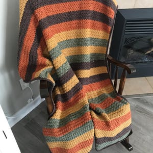 May include: A striped crocheted throw blanket in shades of brown, orange, green, and yellow. The blanket is draped over a wooden rocking chair.