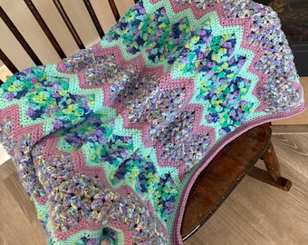 May include: A handmade crochet blanket in shades of purple, teal, and green, draped over a wooden chair. The blanket features a zig-zag pattern with a solid purple border. The yarn has a textured, multi-colored appearance, creating a cozy and inviting aesthetic.