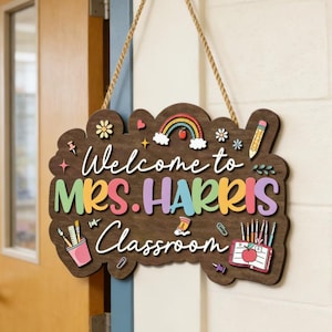 May include: A wooden classroom sign with the text "Welcome to Mrs. Harris' Classroom" in colorful letters. The sign features a rainbow, flowers, pencils, and other school-themed illustrations. It hangs from a rope.