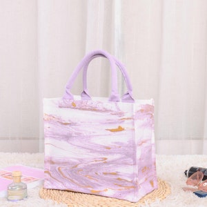Peut inclure: Un sac cabas en toile à motif marbré violet et or sur fond blanc. Le sac a deux anses.
