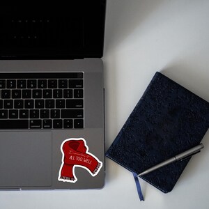 May include: A red scarf with the words "I remember it all too well" printed on it. The scarf is on a laptop computer with a black keyboard and a dark blue notebook with a pen.