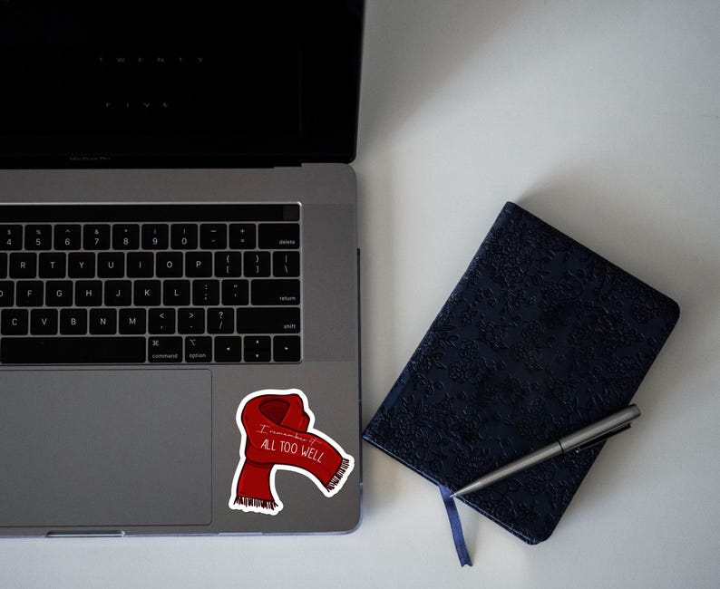 May include: A red scarf with the words "I remember it all too well" printed on it. The scarf is on a laptop computer with a black keyboard and a dark blue notebook with a pen.