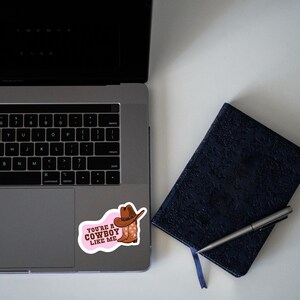 May include: A sticker with the text "YOU'RE A COWBOY LIKE ME" in pink, featuring a cowboy hat and boots. The sticker is placed on a laptop next to a dark blue notebook and a pen.