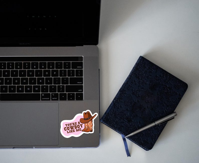 May include: A sticker with the text "YOU'RE A COWBOY LIKE ME" in pink, featuring a cowboy hat and boots. The sticker is placed on a laptop next to a dark blue notebook and a pen.