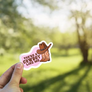 May include: A sticker with a pink and white border featuring a cowboy hat and boots. The text "You're a Cowboy Like Me" is printed on the sticker. The cowboy hat is brown with a silver band, and the boots have a star pattern.