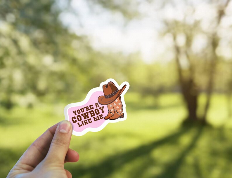 May include: A sticker with a pink and white border featuring a cowboy hat and boots. The text "You're a Cowboy Like Me" is printed on the sticker. The cowboy hat is brown with a silver band, and the boots have a star pattern.