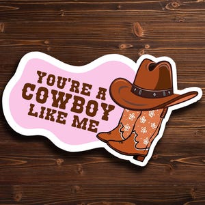 May include: A sticker featuring a brown cowboy hat and boots with a floral design, set against a pink background. The text "YOU'RE A COWBOY LIKE ME" is displayed in brown letters. The sticker is placed on a wooden surface.