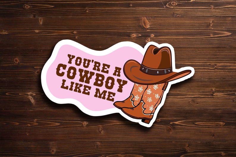 May include: A sticker featuring a brown cowboy hat and boots with a floral design, set against a pink background. The text "YOU'RE A COWBOY LIKE ME" is displayed in brown letters. The sticker is placed on a wooden surface.