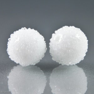 May include: Two white, textured, spherical earrings. Each earring has a bubbly, irregular surface, and they are positioned against a gray background. The earrings are reflected in the surface below.