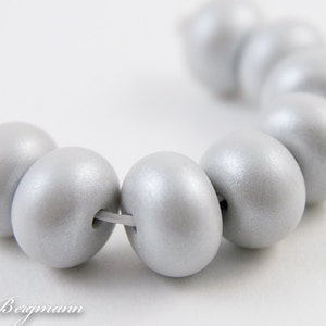 May include: A strand of light gray, oval-shaped beads. Each bead is strung together with a thin, light-colored string. The beads have a smooth, pearlescent finish, reflecting light. The beads are arranged in a curved line.
