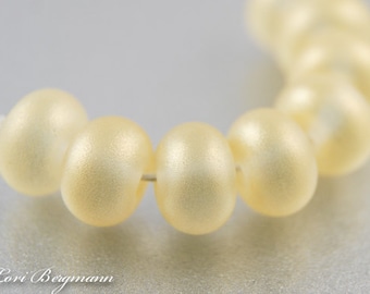 May include: Close-up of a strand of creamy, oval-shaped beads. The beads have a subtle, pearlescent sheen and are strung on a thin, white thread. The beads are evenly spaced and appear to be made of a smooth material.