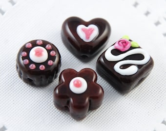 May include: Four dark brown, decorative beads in various shapes: a circle, a heart, a flower, and a square. The circle bead has pink and white accents. The heart bead has a pink heart design. The flower bead has a white and pink center. The square bead has a white swirl and a pink rose.