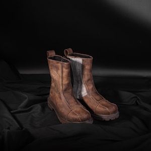 Può includere: Un paio di stivali in pelle marrone con un pannello bianco a contrasto sul retro di ogni stivale. Gli stivali sono su una superficie di tessuto nero.