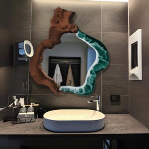 Puede incluir: Un tocador de baño con un lavabo de porcelana blanca y un espejo único de forma irregular. El espejo está enmarcado con madera oscura y resina verde, que se asemeja a una ola rompiendo contra un tronco de árbol.