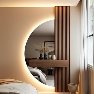 Può includere: Un design moderno per camera da letto con un grande specchio rotondo con una luce calda e brillante. Lo specchio è incastonato in una parete dai toni neutri, completato da un pannello in legno e un piccolo ripiano con vasi decorativi. Un letto è visibile nel riflesso.