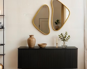 May include: A black dresser with a ribbed front, a vase, a bowl, and two gold-framed mirrors with organic shapes hanging on a white wall.