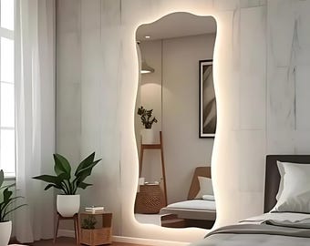 Puede incluir: Un espejo de cuerpo entero, con bordes ondulados, con una luz LED blanca alrededor del perímetro. El espejo está montado en una pared blanca en un dormitorio.