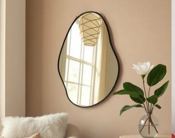 May include: A modern, abstract-shaped wall mirror with a black frame. The mirror reflects a window with white curtains and a light fixture.