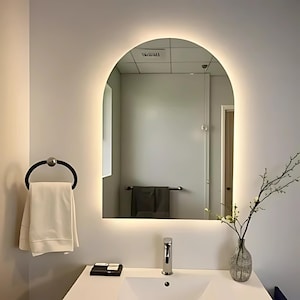 Peut inclure: Une vasque de salle de bain blanche avec un lavabo en porcelaine blanche et un robinet chromé. Une serviette blanche est accrochée à un anneau de serviette noir. Un miroir arqué avec une lumière LED blanche autour du périmètre est monté sur le mur au-dessus du lavabo. Un vase avec une branche de fleurs blanches est posé sur le comptoir.