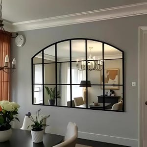 May include: A large arched mirror with a black metal frame divided into 12 squares. The mirror reflects a room with a chandelier, furniture, and plants.