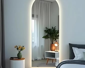 Puede incluir: Un espejo alto y arqueado con iluminación LED integrada. El espejo está enmarcado por una luz suave y brillante, creando una estética moderna. El reflejo muestra un dormitorio con una mesita de noche blanca, una planta y una cama con ropa de cama gris.