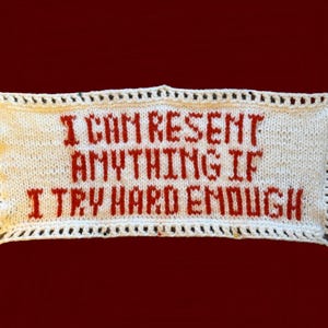 Könnte beinhalten: Rechteckiges, cremefarbenes Strickbanner mit dem Text "I CAN RESENT ANYTHING IF I TRY HARD ENOUGH" in roten Buchstaben. Das Banner hat einen dekorativen Rand mit schwarzen Nähten, vor einem dunkelroten Hintergrund.