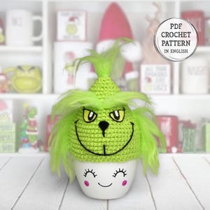 May include: A green crocheted Grinch-themed hat with a matching face on a white ceramic pot. The pot has a smiling face with closed eyes and pink cheeks. The hat has a mischievous expression. The image includes a "PDF Crochet Pattern in English" label.