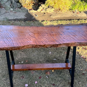 Puede incluir: Una mesa de madera de borde vivo con un acabado de madera natural y patas de metal negro. La mesa tiene un estante debajo.