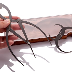 May include: Two metal drawing compasses with adjustable arms and points, in a wooden box with a clear lid.