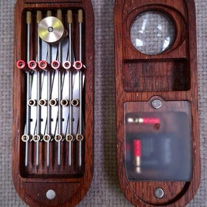May include: A two-part wooden case with a dark brown finish. The left side holds five parallel metal needles with red and gold accents and a circular brass knob. The right side features a circular magnifying lens and a translucent compartment.
