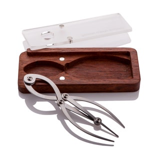 May include: A silver compass tool with a wooden case. The compass has two arms with a ball at the end of each arm. The case is made of dark wood and has a clear plastic lid.