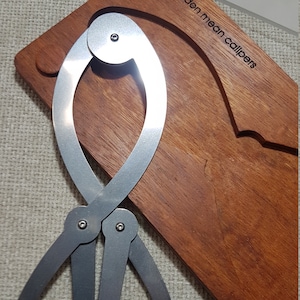 May include: A silver golden mean calipers tool on a wooden base with the text "golden mean calipers" on the base.