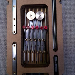 Op de afbeelding: Een rechthoekig bruin houten opbergplateau met een metalen tekeninstrument met meerdere punten, twee lange dunne metalen staven, twee zilverkleurige metalen strips met schroefgaten en een zwarte adapter met rode knoppen.