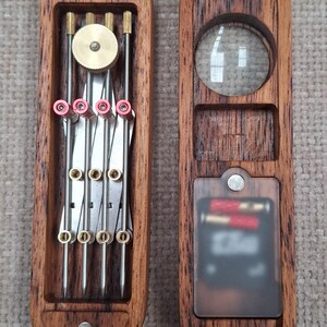 May include: A two-piece wooden case with a dark wood grain finish. The left side holds a metal drafting compass tool with brass and red accents. The right side features a circular magnifying lens and a rectangular compartment with a translucent cover.