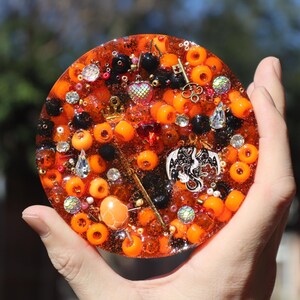 Op de afbeelding: Een ronde, oranje hars coaster gevuld met oranje en zwarte kralen, edelstenen en bedels. Bedels zijn onder meer een sleutel, een kroon, een hart en een draak. De coaster wordt in een hand gehouden.