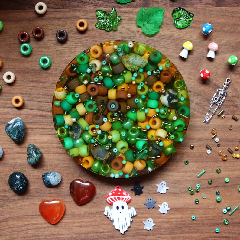 May include: A circular resin piece filled with green, yellow, and brown beads, surrounded by decorative elements. The arrangement includes gemstones, leaf-shaped charms, miniature mushrooms, and a skeleton charm, all on a wooden surface.
