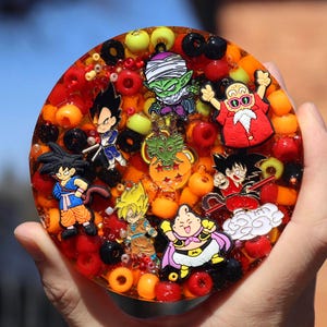 Puede incluir: Un posavasos redondo de resina naranja con cuentas de colores y varios amuletos de personajes de anime. Los amuletos incluyen un dragón, un personaje con una camisa roja, un personaje con una camisa verde y un personaje con una camisa azul.